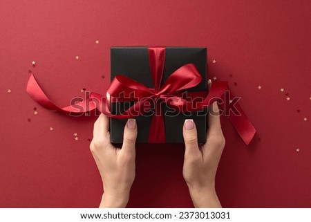 Dive into holiday magic! First person top view of hands reach for lavish black giftbox featuring ribbon bow, nestled among shimmering confetti on marsala backdrop. Perfect for Black Friday marketing
