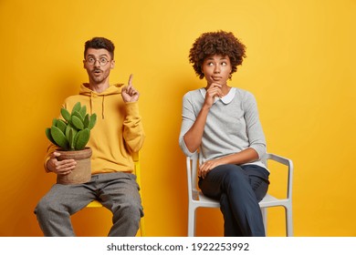 Diverese woman and man sit separately on chairs wait in queue isolated over yellow background. Surprised guy in sweatshirt holds cactus points above tells about something amazing poses near wife