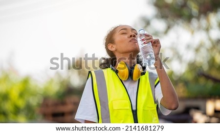 Divergent A African american woman foreman or worker wearing Cross Eye safety is drinking a bottle of water after finishing work and relaxing on the old truck at cargo container port