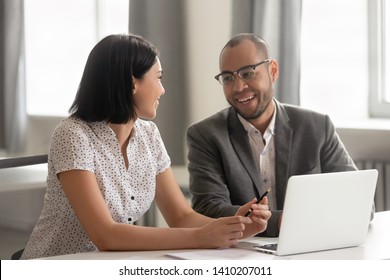 Diverse African American And Asian Happy Colleagues Talking Laughing Working Sharing Ideas Together In Office At Work Break, Smiling Coworkers Having Friendly Conversation Sitting At Desk With Laptop