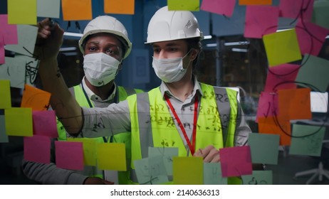 Diverse architects in safety mask and hardhat discuss and write on glass board with sticky notes working together in office - Powered by Shutterstock