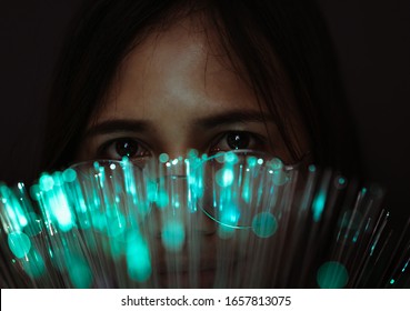 Diverse Asian Girl Looks Through Futuristic Teal Coloured Bokeh - Millennial Woman Looking At Camera With Advanced Fibre Optic Technology - Next Generation Super Fast Internet And Optical Tech Concept