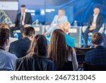 A diverse audience attentively listens to speakers on stage during a business conference. The panelists appear out of focus as they discuss key topics.