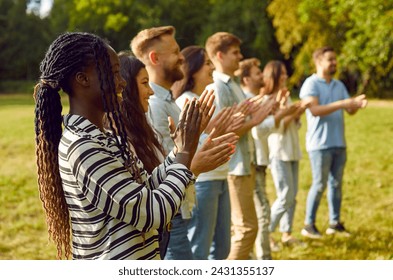 Diverse audience enjoying outdoor summer event such as music festival or community fair. Happy young Caucasian and African American people standing in park, looking away, clapping hands and smiling - Powered by Shutterstock