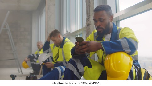Diverse builders at construction site resting during break using smartphone. Multiethnic workers relaxing on floor scrolling mobile phone on break doing houses renovation