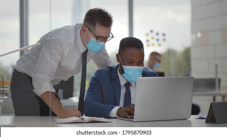 Diverse Business Colleagues In Protective Mask At Office Desk Working On Laptop. Caucasian Employee In Facial Mask Helping African Coworker With Business Project In Office