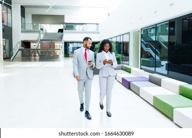 Diverse Business Professionals Consulting Internet On Mobile Phones. Business Man And Woman Walking Through Office Hallway And Using Smartphones. Wireless Communication Concept