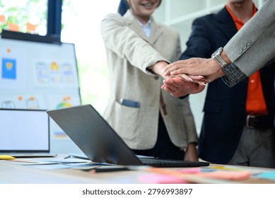 Diverse business professionals stack hands over a desk with laptop and documents, symbolizing teamwork, collaboration, and partnership for corporate use.