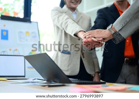 Diverse business professionals stack hands over a desk with laptop and documents, symbolizing teamwork, collaboration, and partnership for corporate use.