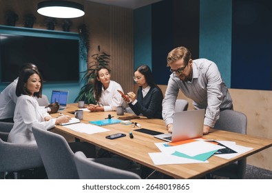 Diverse business team discussing project strategy, young man leaning over laptop while colleagues collaborate using smartphones and digital tools in modern meeting room