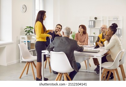 Diverse business team having a work meeting in a modern office. Group of happy multiethnic people sitting around a table with laptops and tablets and listening to a young woman making a suggestion - Shutterstock ID 2112104141