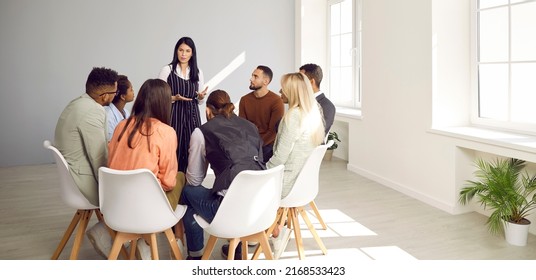 Diverse Business Team Meeting In Modern White Office. Group Of People Listening To Serious Young Woman Who's Standing In Front Of Them, Explaining Some Rules And Talking About Importance Of Management