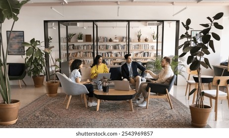 Diverse business team talking in modern co-working meeting space, sitting on couch, armchairs, using laptops, discussing work communication, teamwork, startup project strategy - Powered by Shutterstock