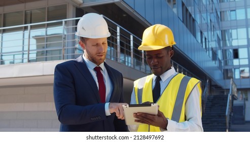 Diverse Businessman And Architect In Hardhats With Tablet Pc Computer At Construction Site. Multiethnic Entrepreneur And Engineer Using Tablet Pc Standing Outside Modern Building