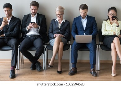 Diverse businesspeople applicants or customers sitting in row using devices phones and laptop, multi ethnic men women waiting for job interview in queue, human resources, people and gadgets concept