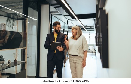 Diverse businesspeople sharing a laugh during a discussion in an office. Two happy business colleagues using a digital tablet while walking together in a modern workspace. - Powered by Shutterstock