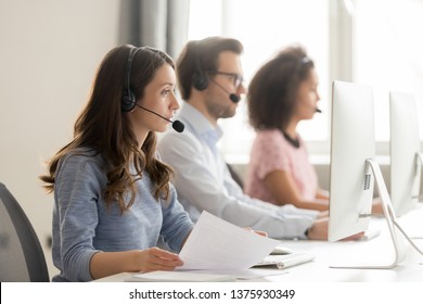 Diverse call center workers sitting in row, focus on customer service member girl in headset looks at pc screen hold paper talk with potential client provide insurance information sell company product