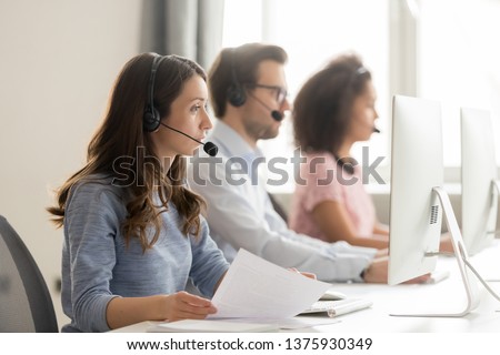 Diverse call center workers sitting in row, focus on customer service member girl in headset looks at pc screen hold paper talk with potential client provide insurance information sell company product