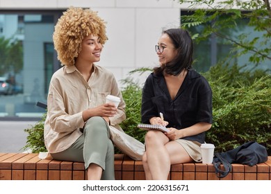 Diverse cheerful young women have pleasant conversation take interview write answers to questionnaire drinks takeaway coffee dressed in casual clothes pose on bench against urban background. - Powered by Shutterstock