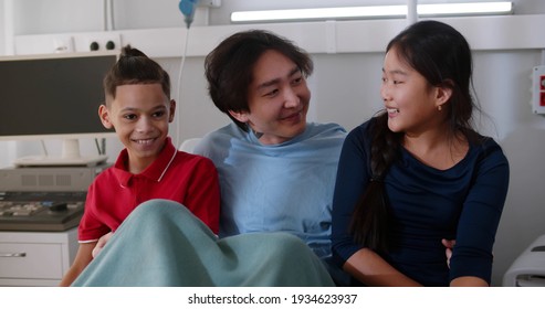 Diverse Children Visiting Adoptive Asian Father In Hospital Ward. Sick Korean Man Resting In Hospital Bed And Hugging Kids Visiting In Clinic Room. Healthcare And Family Concept