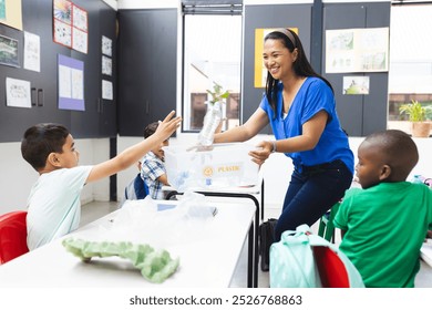 A diverse classroom learns about recycling. Middle-aged Asian teacher smiles, young biracial boy offers plastic bottle, young African American boy observes, surrounded by posters, learning, education, - Powered by Shutterstock