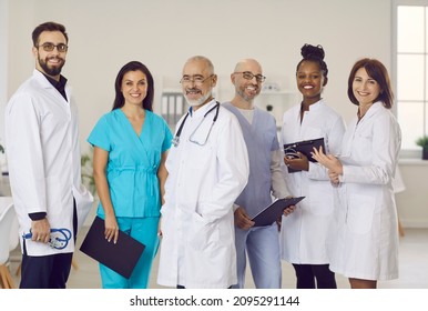 Diverse clinic staff at work. Team of happy young and mature doctors, clinicians, therapists, cardiologists in scrubs and white coat uniforms standing in office, holding clipboards, looking at camera