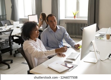 Diverse Colleagues Discussing Online Work Together, Female African American Intern Ask Questions To Mentor, Looking At Screen, Executive Boss Helping Employee With Computer Work In Open Office Space