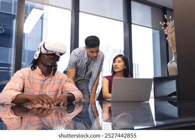 Diverse colleagues explore virtual reality and use laptop in a modern office. An African American man wears a VR headset, Asian woman and a biracial man look at laptop screen, unaltered - Powered by Shutterstock