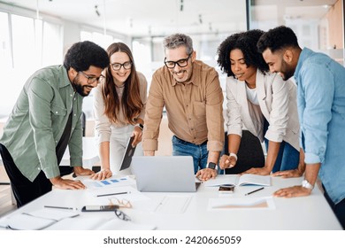 Diverse colleagues gather around a laptop with bright expressions, indicating a successful collaborative effort and a lively brainstorming session in a well-lit, modern workspace, team work concept - Powered by Shutterstock