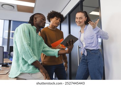 Diverse colleagues share content on tablet in a modern office. An African American man with short hair, a biracial man with curly hair, and a Caucasian woman with long hair laugh together - Powered by Shutterstock