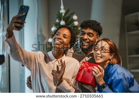 Diverse colleagues are smiling while taking a group selfie with a smart phone, celebrating a christmas party at the office, with one person holding a red gift box