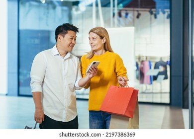 Diverse Couple Asian Man And Blonde Woman In Clothes Supermarket, Looking At Mobile Phone Screen, Choosing Shopping, Holding Colorful Shopping Bags And Gifts.