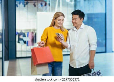 Diverse Couple Asian Man And Blonde Woman In Clothes Supermarket, Looking At Mobile Phone Screen, Choosing Shopping, Holding Colorful Shopping Bags And Gifts.