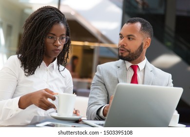 Diverse Couple Of Colleagues Watching Project Presentation On Laptop. Business Man And Woman Drinking Coffee, Using Computer, Looking At Screen And Talking. Teamwork Concept