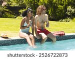 Diverse couple enjoys drinks by the poolside, laughter evident in their sunny disposition. She wears a striped swimsuit and he in red shorts, both relishing a bright, leisurely day outdoors.