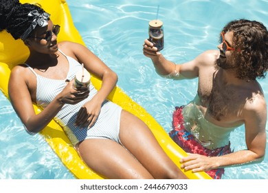 A diverse couple enjoys drinks in pool. The African American woman, with a headwrap, and Caucasian man, bearded, toast on floaties, unaltered - Powered by Shutterstock