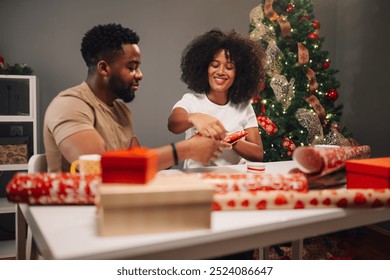 Diverse couple is filled with joy, wrapping Christmas gifts in red paper, surrounded by a festively decorated tree. The image captures a cozy and cheerful atmosphere. - Powered by Shutterstock