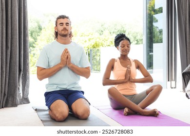 A diverse couple finds peace in yoga at home. A Caucasian man and African American woman, eyes closed, hands together, dressed for exercise, unaltered - Powered by Shutterstock