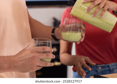 Diverse couple pouring green smoothies into glasses. Modern kitchen with blender on counter, white cabinets, and stainless steel appliances, unaltered - Powered by Shutterstock