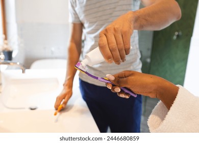 A diverse couple is preparing for their dental hygiene routine. The African American woman and her Caucasian partner are squeezing toothpaste onto brushes, ready to brush their teeth, unaltered - Powered by Shutterstock