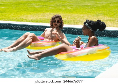 A diverse couple relaxes on pool floats, enjoying drinks at home. The Caucasian man and African American woman sport swimwear and sunglasses in sunshine, unaltered - Powered by Shutterstock