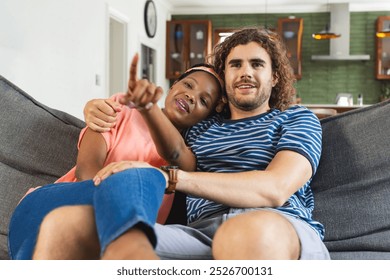 A diverse couple relaxes on sofa at home, pointing at something while watching TV. She is African American with black hair; he is Caucasian with curly brown hair, unaltered. - Powered by Shutterstock