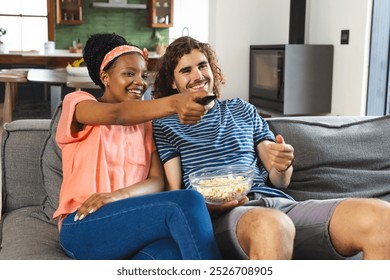 A diverse couple relaxes on sofa, sharing popcorn. She points at the TV; he smiles with the remote, in warm home, unaltered - Powered by Shutterstock