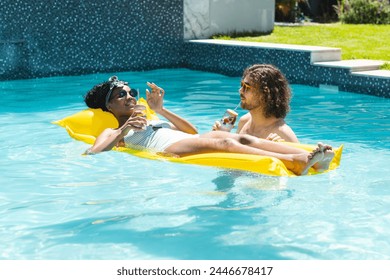 A diverse couple relaxes on yellow pool float. The African American woman and Caucasian man enjoy drinks, basking in sun with sunglasses, unaltered - Powered by Shutterstock