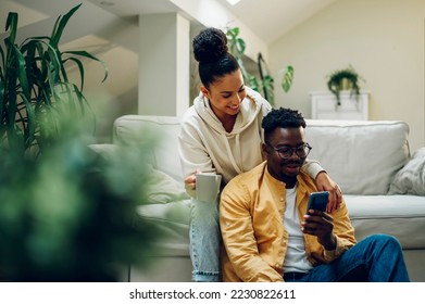 Diverse couple relaxing on sofa at home and drinking coffee while using smartphone. African american man and hispanic woman sitting on couch while chatting and talking. Copy space. Online shopping. - Powered by Shutterstock