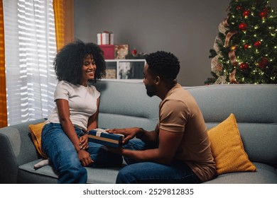 A diverse couple shares a cheerful moment exchanging a small gift while sitting on the sofa, surrounded by the glow of the holiday season and lively Christmas decorations. - Powered by Shutterstock