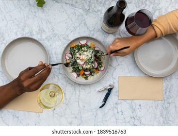 A diverse couple shares a glass of wine and a plate of greens while dining outside at a beautiful white marble table. 