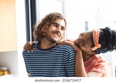 Diverse couple shares a playful moment in a bright kitchen setting. The young woman with an orange headband smiles at the man with curly hair, creating a warm, affectionate atmosphere. - Powered by Shutterstock