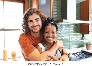 A diverse couple shares a warm embrace in a modern kitchen at home. The Caucasian man has curly hair, while the African American woman sports a radiant smile with a headband, unaltered. - Powered by Shutterstock