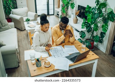 Diverse couple sitting at home office with a laptop at the table at home and looking the blueprints of their new house. Investing money into new home. Making financial strategy. Copy space. - Powered by Shutterstock